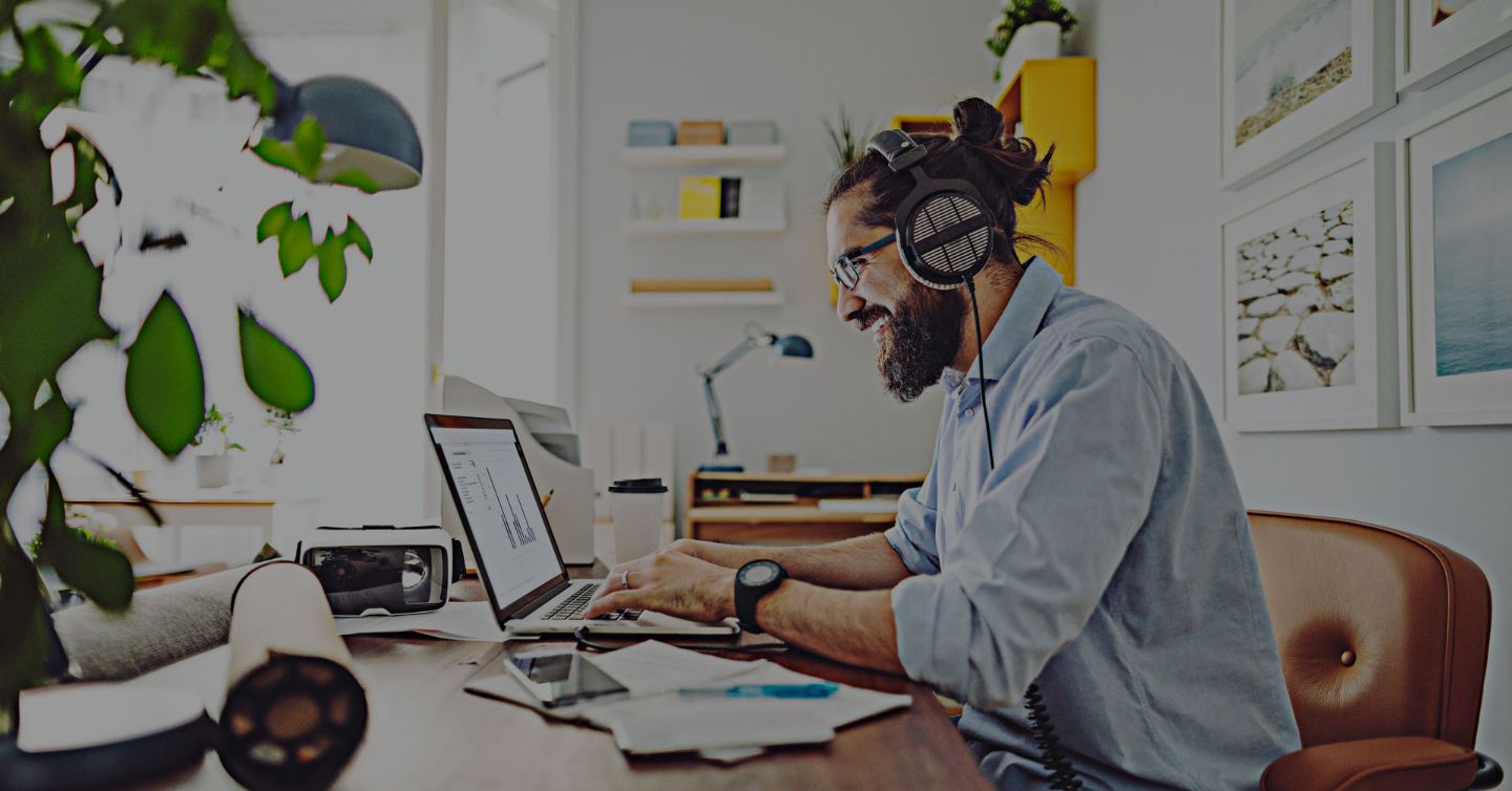 Man in a shirt sitting at a desk working on a laptop with headphones on